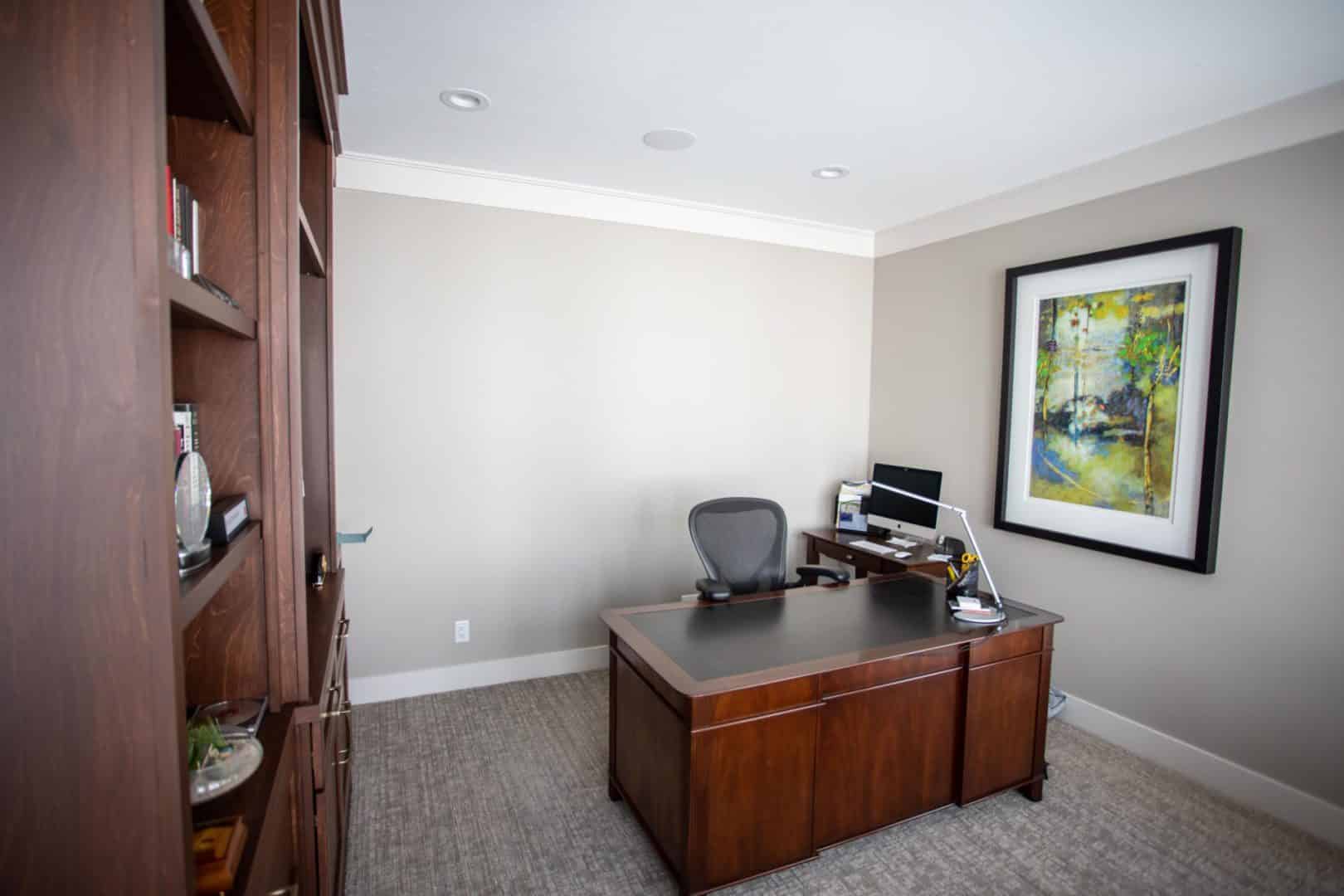 Office space with wooden desk, ergonomic chair, and colorful landscape painting.