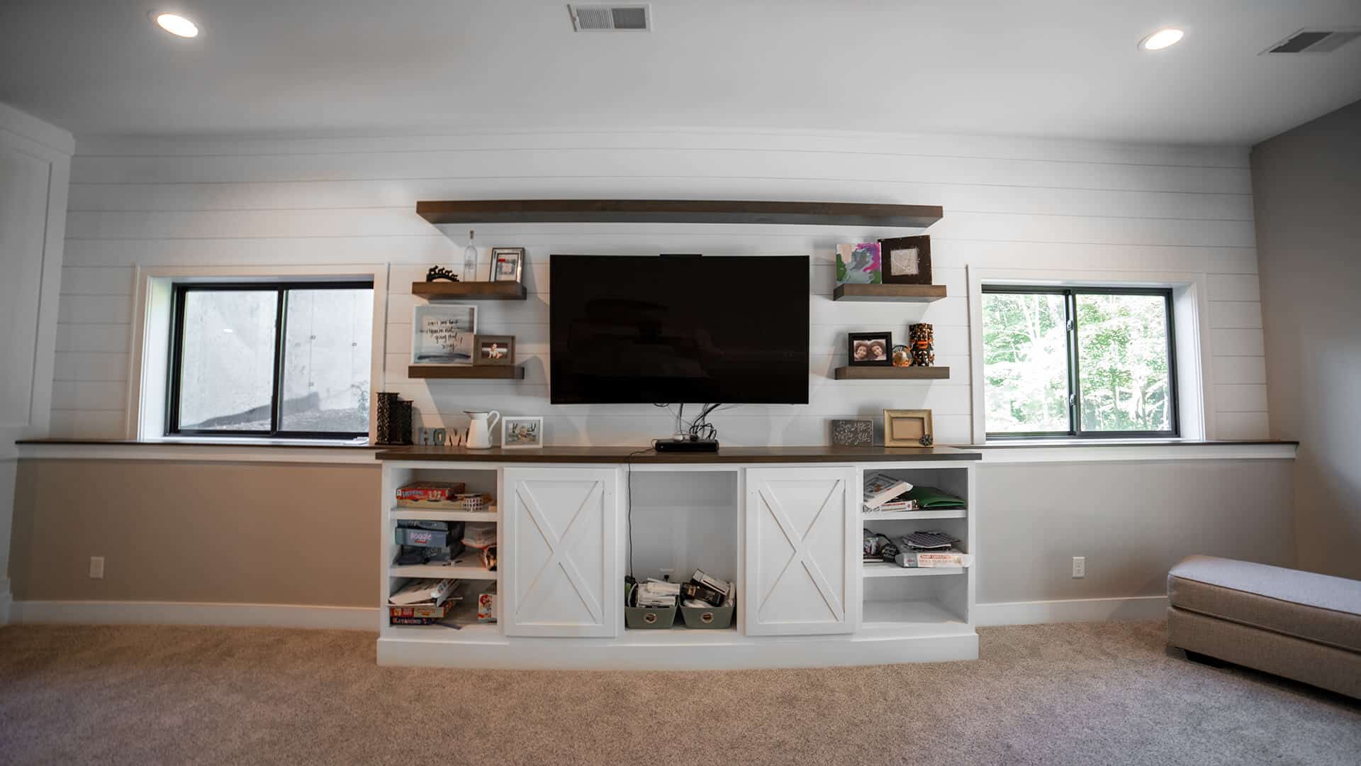 Modern living room with built-in shelves and large TV, bright windows, and neutral decor.