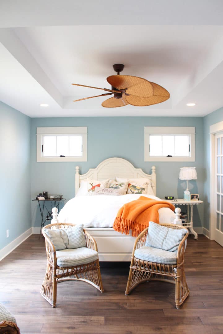 Coastal bedroom featuring a rattan ceiling fan, light blue walls, and a cozy bed with decorative pil.