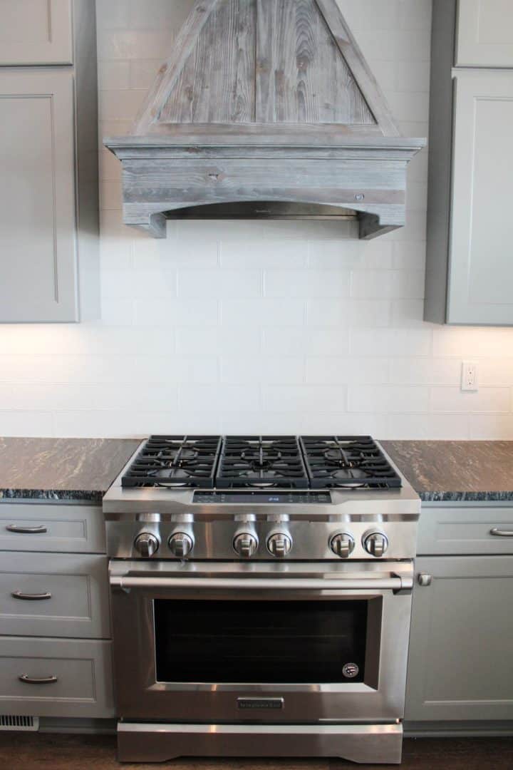 Stainless steel kitchen stove with gray cabinets and rustic wooden range hood.