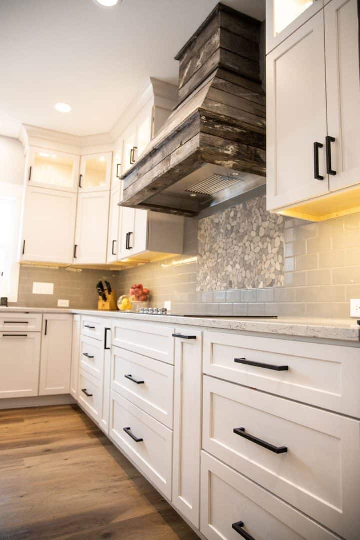 Modern white kitchen with wooden range hood, gray backsplash, and black cabinet handles.