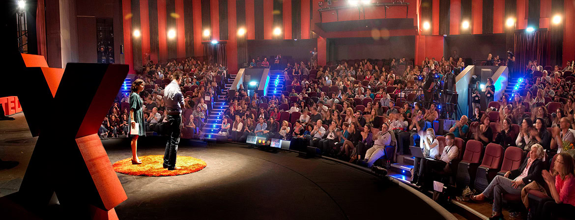 A large audience seated in a theatre-style venue with a stage and bright lighting.