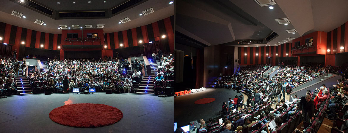 A large audience seated in a theatre-style auditorium with a red circular carpet on the stage.