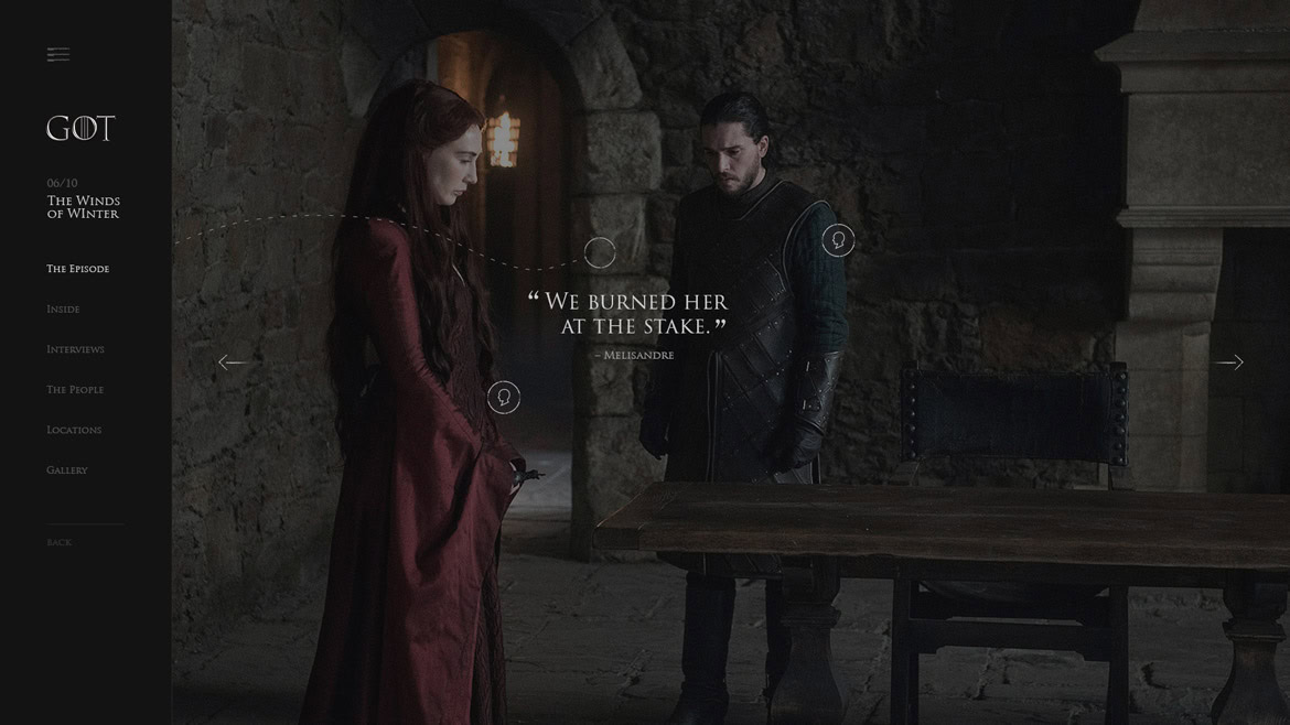 A woman in a red dress and a man in dark clothing stand in a stone-walled room with a wooden table.