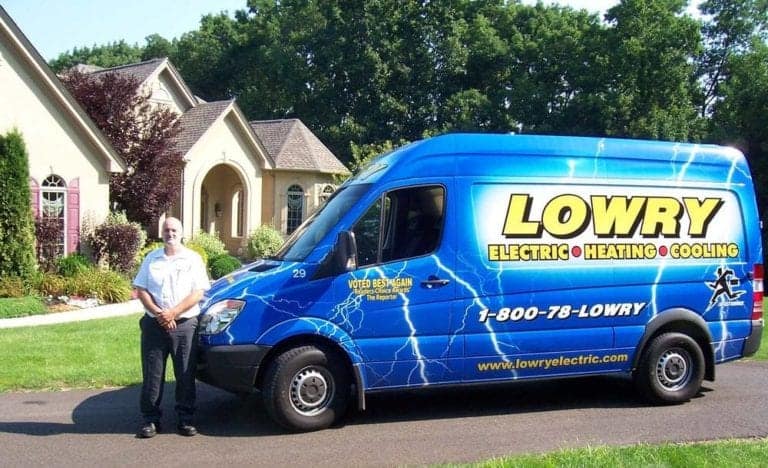 A man in uniform stands next to a blue Lowry Electric, Heating & Cooling service van parked in a residential driveway, ready to assist customers throughout the local service area.