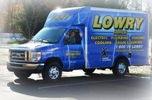 A blue Lowry Services truck with lightning graphics advertises electric, plumbing, heating, cooling, and drain cleaning services—plus details about their maintenance plan—parked in a lot.