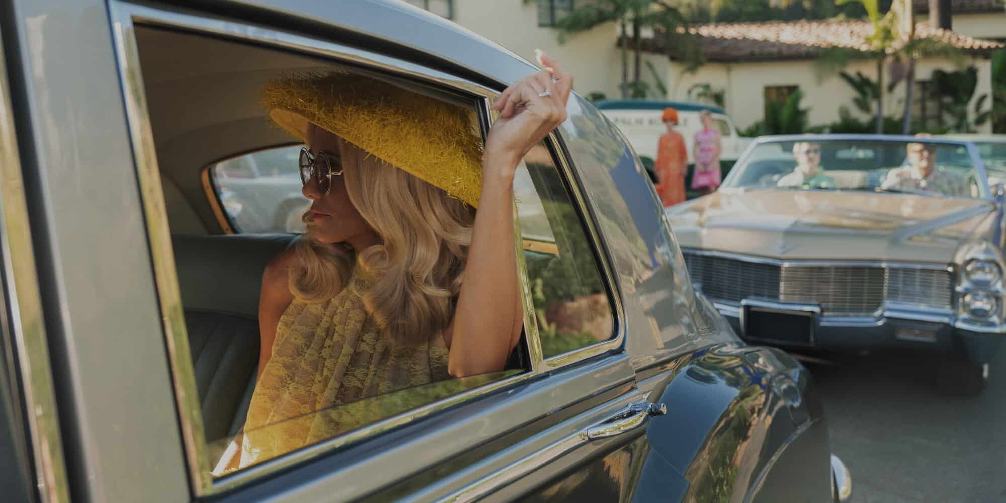 Elegant kvinna i vintageoutfit och solglasögon i en klassisk bil med bredbrättad solhatt och transparent lockigt hår, under en solig dag.
