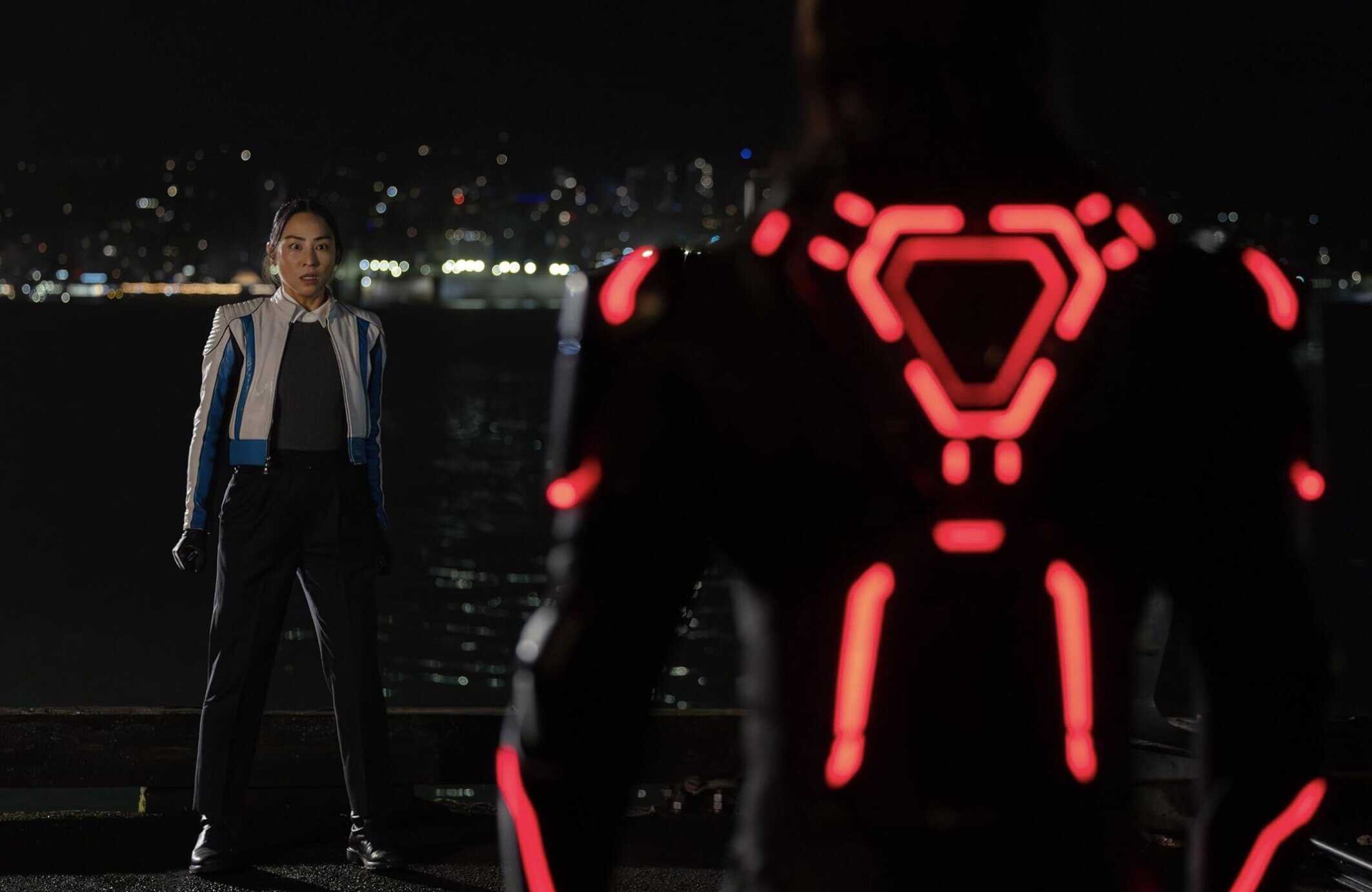 Mångfärgad sci-fi robot i neonljus med futuristisk design står inför en kvinnlig figur vid nattstid vid vattnet med stadens ljus i bakgrunden, vilket ger en modern, futuristisk och cyberpunk atmosfär.