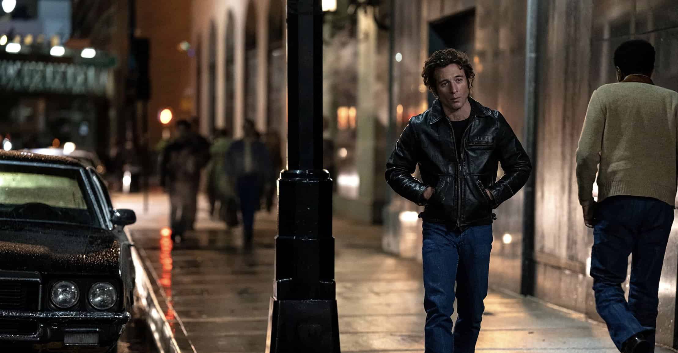 Mannen i svart skinnjacka på stadsgata på kvällen, med neonljus och regndroppar som reflekteras i gatubelysningen, vilket ger ett urban och stilrent intryck.