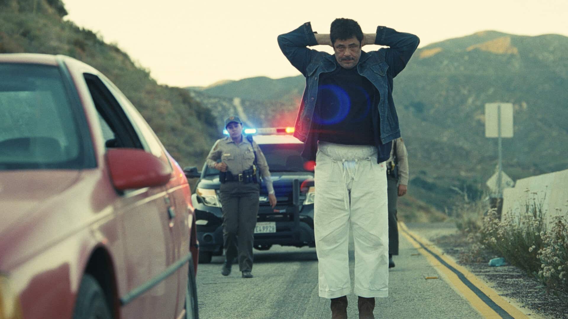 1. En man står på vägbanan med händerna bakom huvudet medan poliser undersöker ett trafikstopp, med en röd bil i förgrunden och berg i bakgrunden.