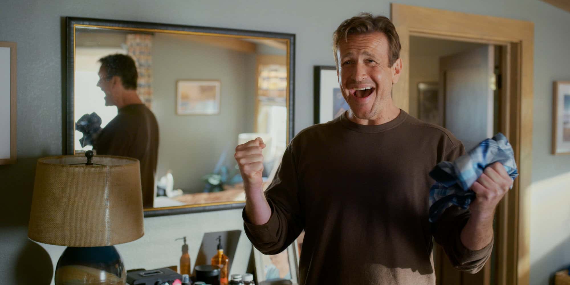 Upprymd man med skjorta i hand, visar glädje över att få tag på kläder, i ett mysigt hem.