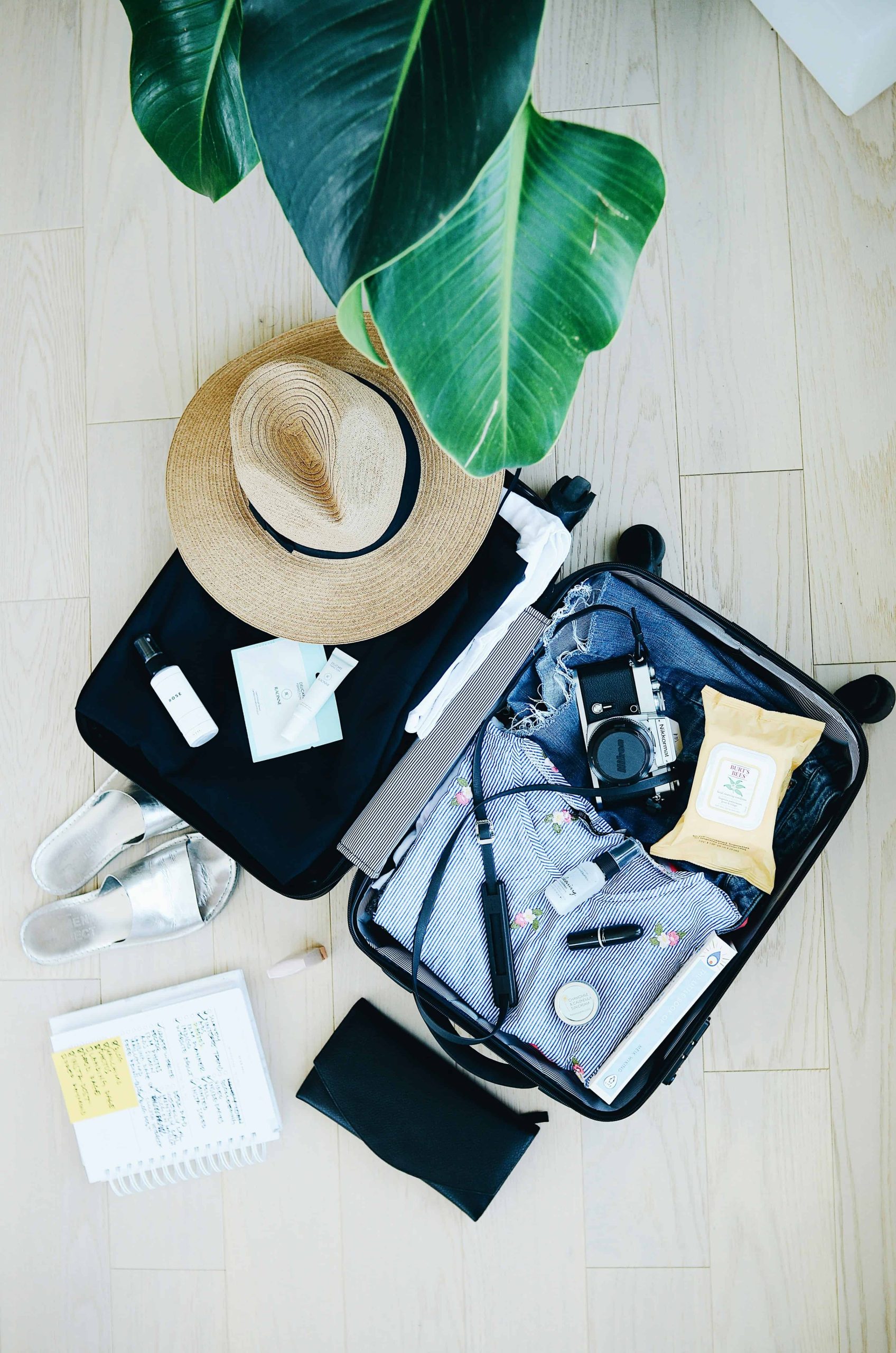 Luxury travel packing essentials in an open suitcase with a straw hat and tropical leaves.