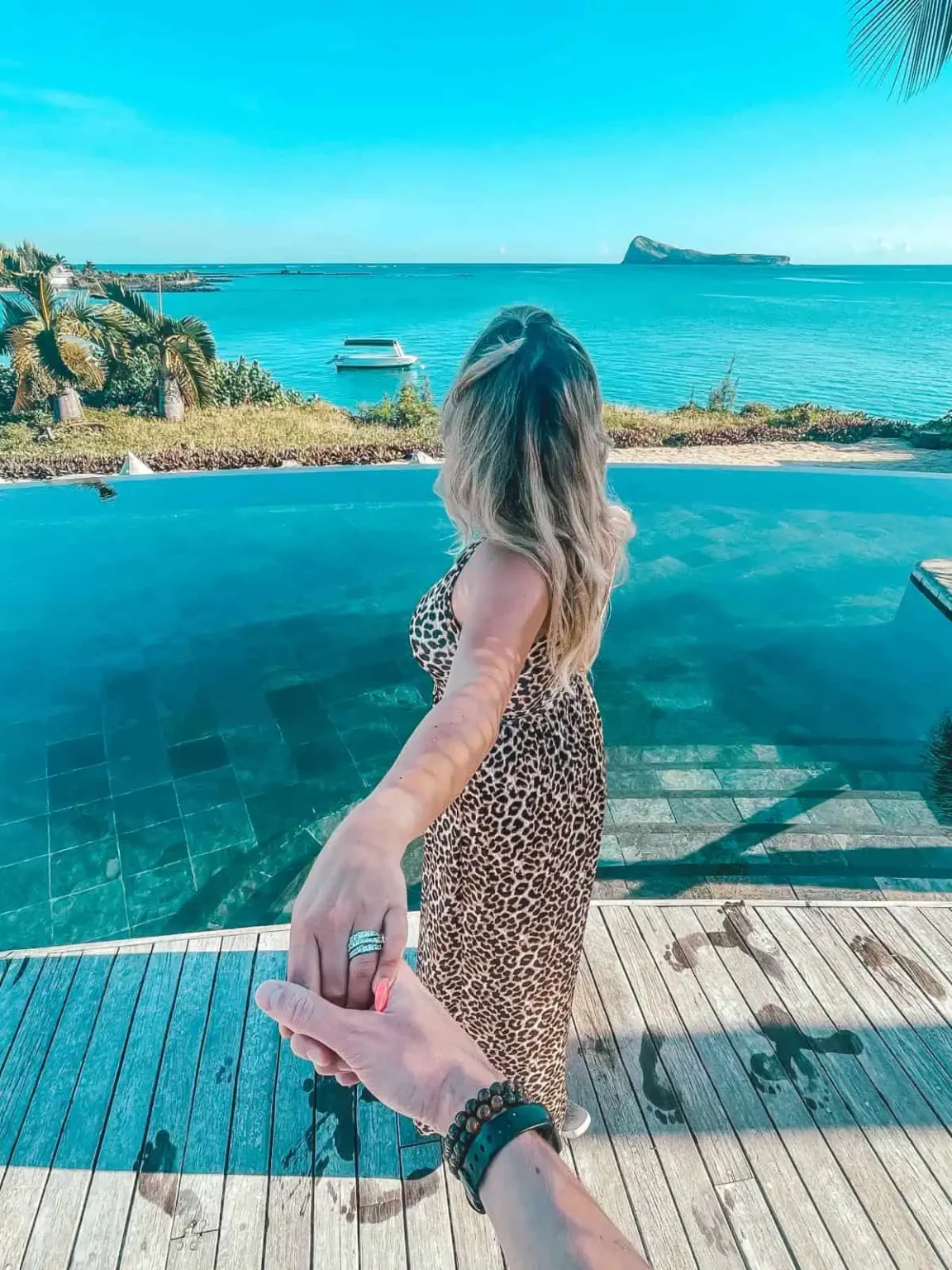 Woman in leopard print dress holding hand by infinity pool overlooking ocean.
