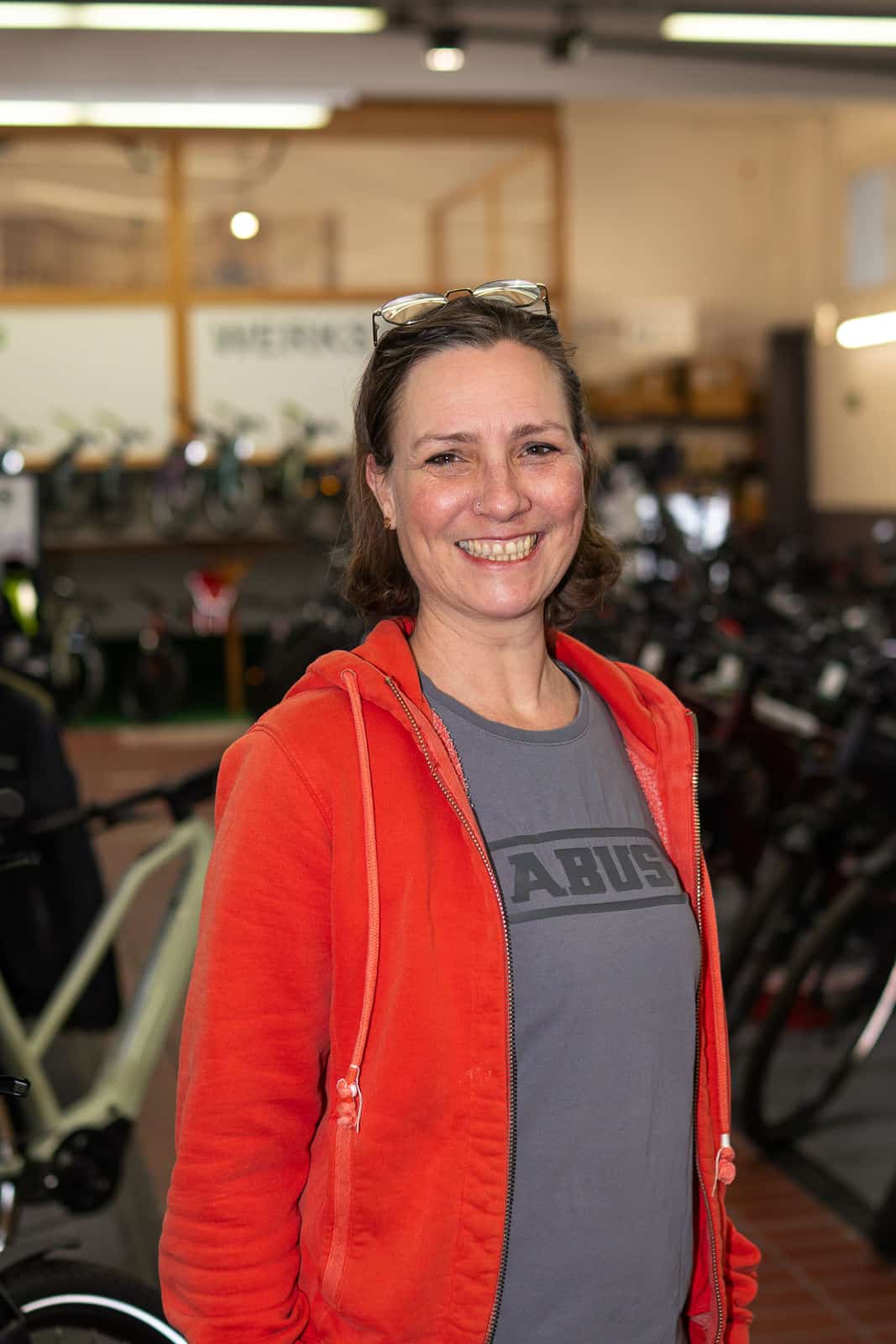 5 - 260128_shotitt-Monz-Fahrradwelten-Portraits-05_Johanna Wolf Lächelnde Frau in Fahrradgeschäft mit roten Jacke und Brille auf Kopf.