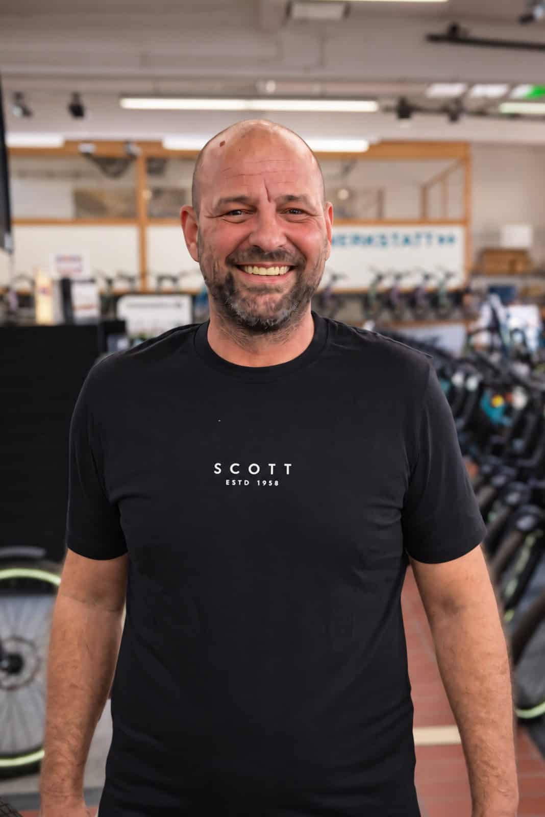 MONZ Fahrradwelten Mitarbeiter Dennis Melzer Lächelnder Mann in Fitnessstudio mit schwarzen T-Shirt.