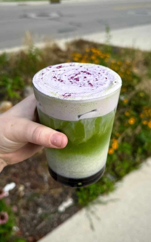 Vegan matcha latte topped with purple drizzle, served in a clear glass outdoors.