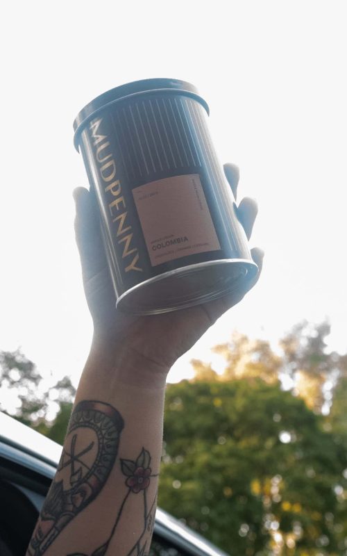 Mudpenny coffee container held up outdoors with trees in the background.