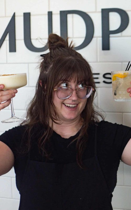 Woman holding two cocktails at Mudpenny bar, smiling and enjoying drinks.