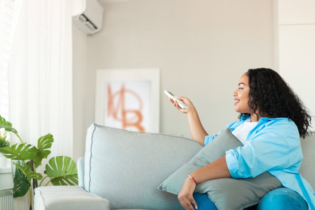 COMFORTABLE WOMAN RELAXING AT HOME, CONTROLLING HVAC SYSTEM WITH REMOTE, SHOWCASING TOP-RATED HVAC AND PLUMBING SERVICES FOR HOME COMFORT.