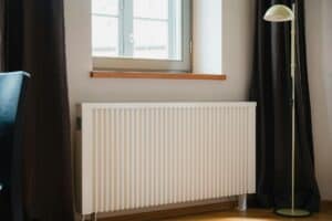 EFFICIENT HOME HEATING RADIATOR UNDER WINDOW WITH BLACK CURTAINS AND FLOOR LAMP, SHOWCASING PROFESSIONAL HVAC SERVICES FOR OPTIMAL INDOOR CLIMATE CONTROL.