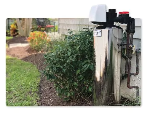 A stainless steel water softener unit is installed outside next to a house, surrounded by plants and mulch. For installation details or questions, please contact us.