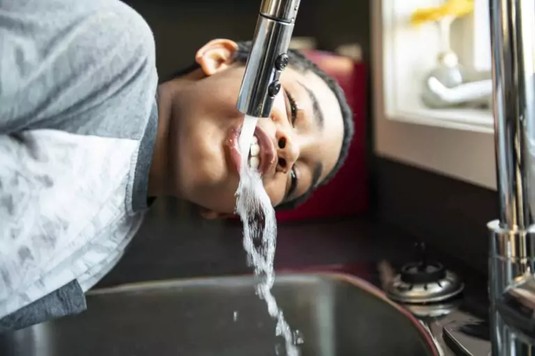 A boy leans over a kitchen sink, drinking water directly from a running faucet, his face in close contact with the cool stream.