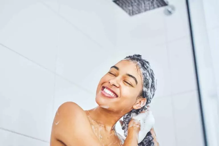 A person is smiling while washing their hair with shampoo under a shower in a tiled bathroom, enjoying the soothing contact of water and suds.
