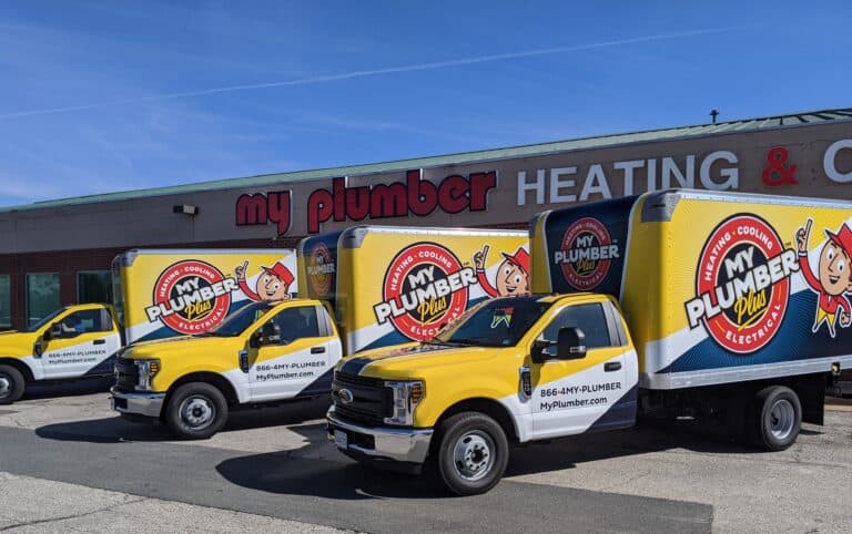 Three yellow and white My Plumber vans are parked outside a building with a "my plumber" sign, "HEATING & COOLING" lettering, and expert Drain Services available for all your plumbing needs.