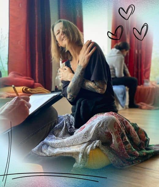 Eine Frau mit Tätowierungen sitzt auf dem Boden, umarmt sich und lächelt, während eine andere Person in der Nähe auf einem Klemmbrett schreibt; im Hintergrund sitzen zwei Personen, und in der rechten oberen Ecke sind drei gezeichnete Herzen zu sehen.