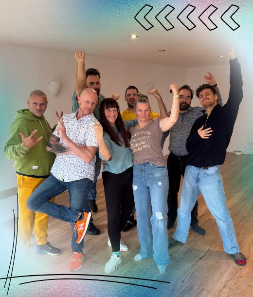 Eine Gruppe von acht Erwachsenen, einschließlich ihres Ausbilders, posiert zusammen in einem Innenraum, lächelnd und mit energischen Gesten, mit einer bunten Umrandung und dekorativen Pfeilen in der oberen rechten Ecke - um den lebendigen Geist ihrer NLP-Ausbildung einzufangen.