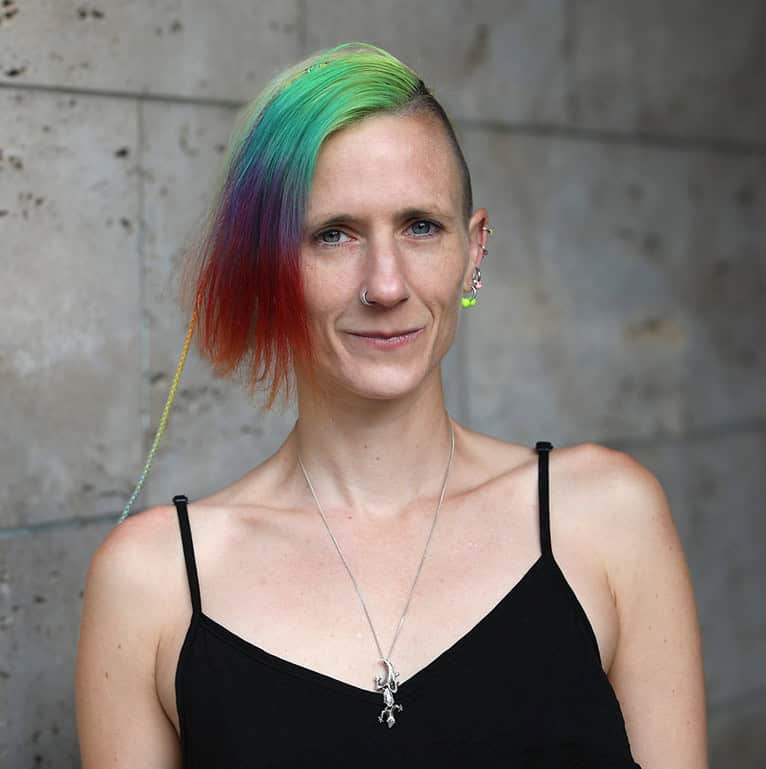 Person mit kurzem regenbogenfarbenem Haar, mehreren Ohrpiercings und einem Nasenring, die ein schwarzes Trägertop und einen silbernen Anhänger trägt und vor einem Steinmauer-Hintergrund steht.