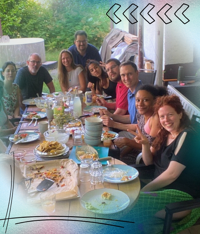 Eine Gruppe von zehn Personen sitzt an einem Tisch im Freien, isst gemeinsam und diskutiert darüber, wie man nlp kostenlos lernen kann. Teller mit Essen und Getränken sind auf dem Tisch ausgebreitet, und jeder lächelt in die Kamera.