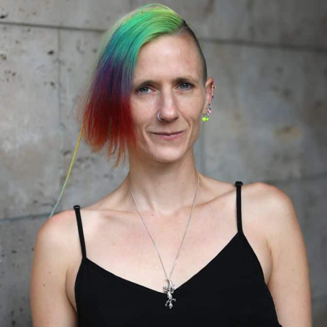 Person mit kurzem, asymmetrischem regenbogenfarbenem Haar, die ein schwarzes Unterhemd, mehrere Ohrringe und eine Halskette trägt und an einer Steinwand steht.