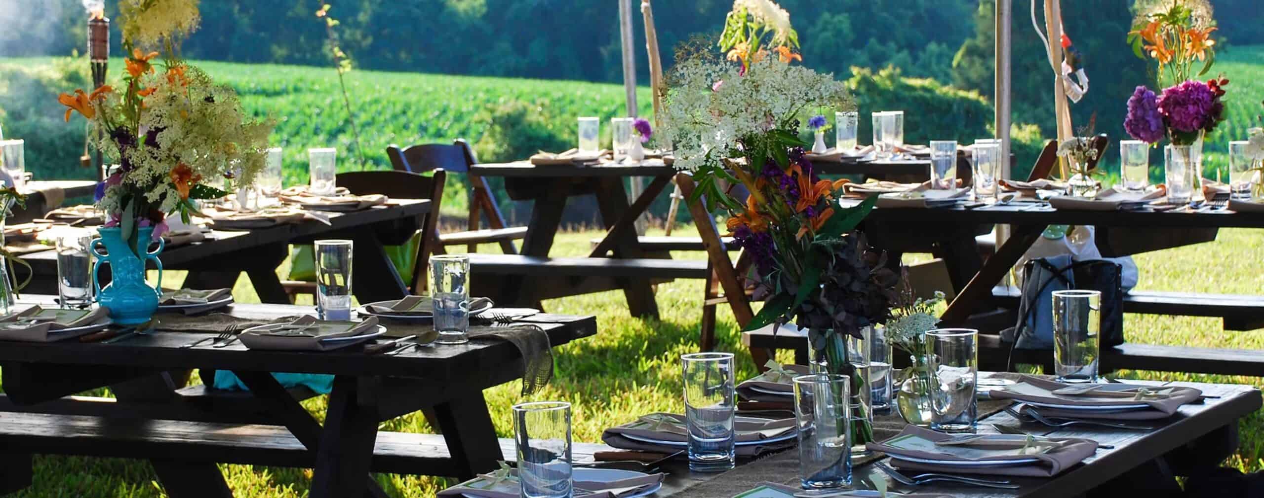 Buitenlunch setup met tafels, bloemen en glazen in een groene omgeving.