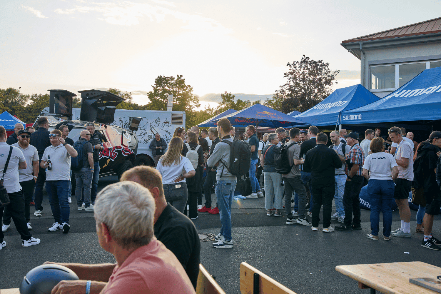 Menschen bei einem Outdoor-Event mit Zelten und Bühnen, Sonnenuntergang im Hintergrund.