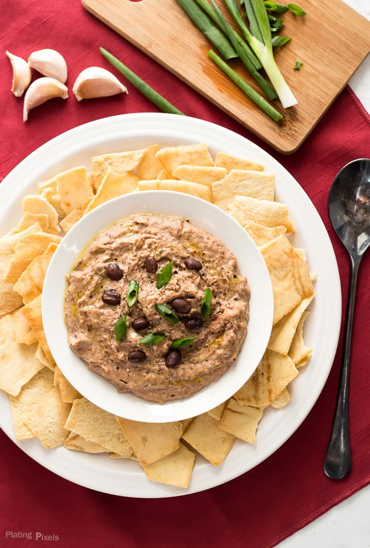 Sun Dried Tomato Black Bean Hummus in a bowl with chips around it and garlic cloves at the side