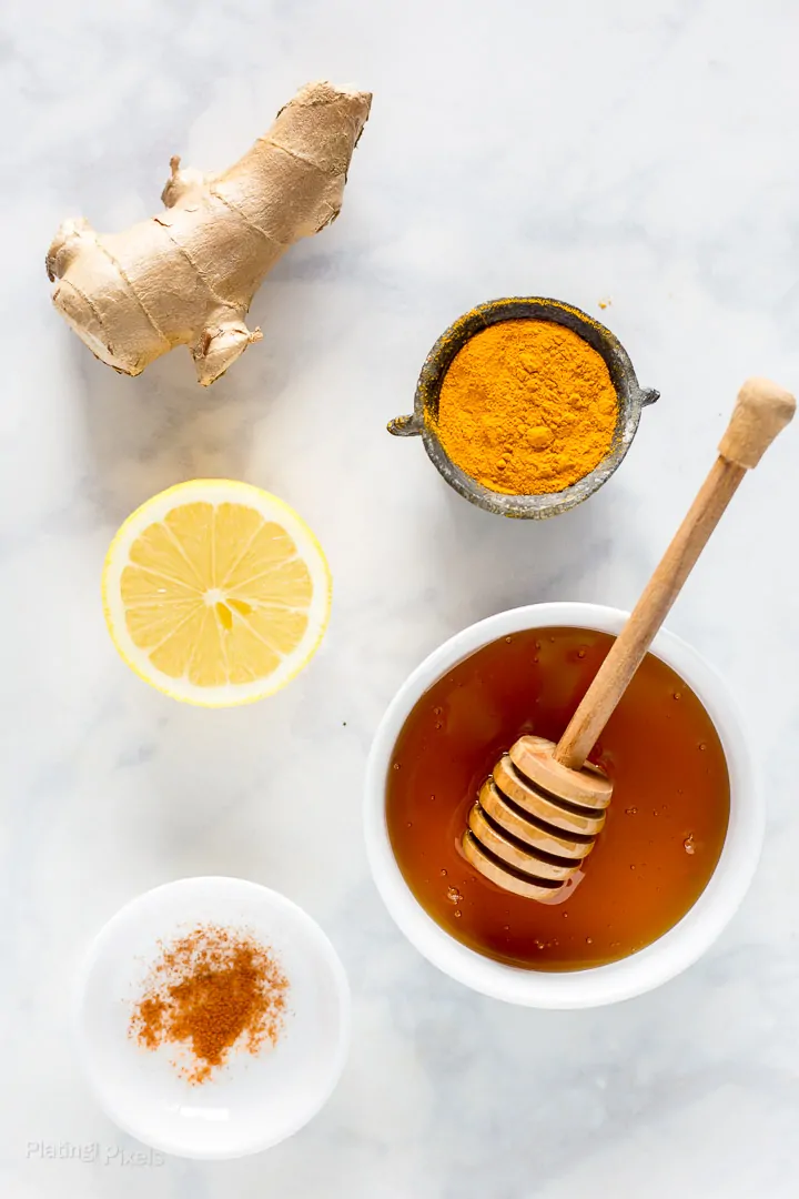 Ingredients to make Turmeric Tea on a marble table, including fresh ginger root, turmeric powder, honey, and cayenne pepper
