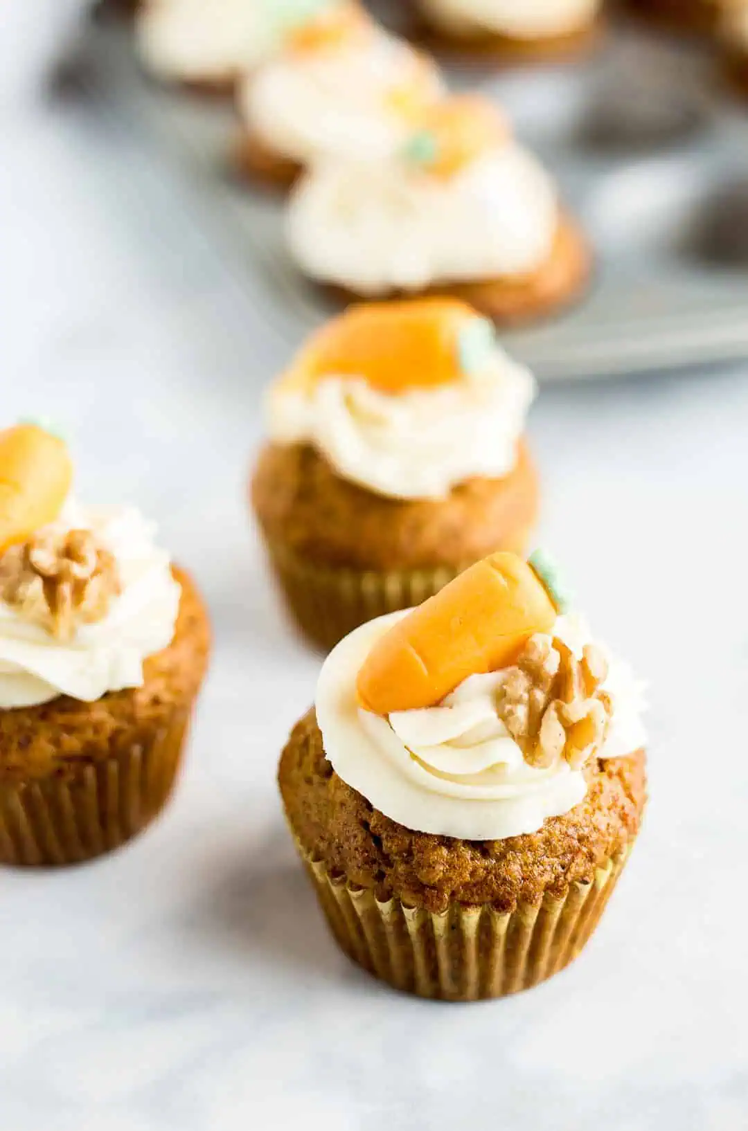 Three frosted and decorated Carrot Cake Cupcakes next to a pan of additional cupcakes