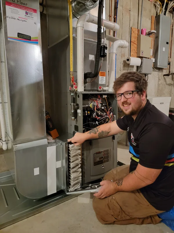 A technician kneels beside an open HVAC unit, pointing to the air filter compartment with tools nearby in a basement setting.