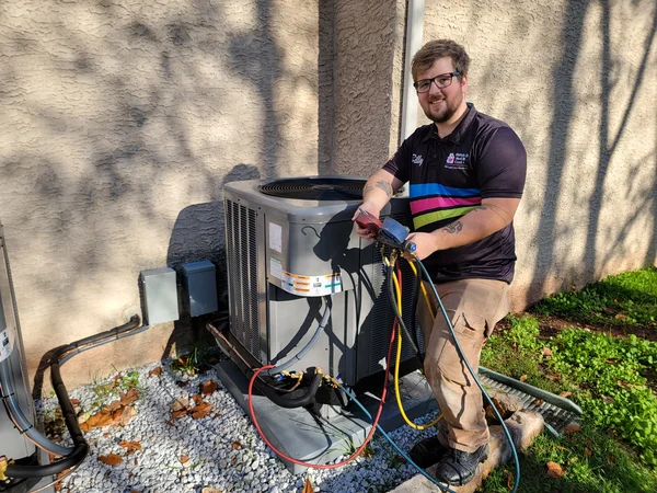 A technician stands next to an outdoor HVAC unit using diagnostic tools, with hoses connected, on a sunny day beside a house.