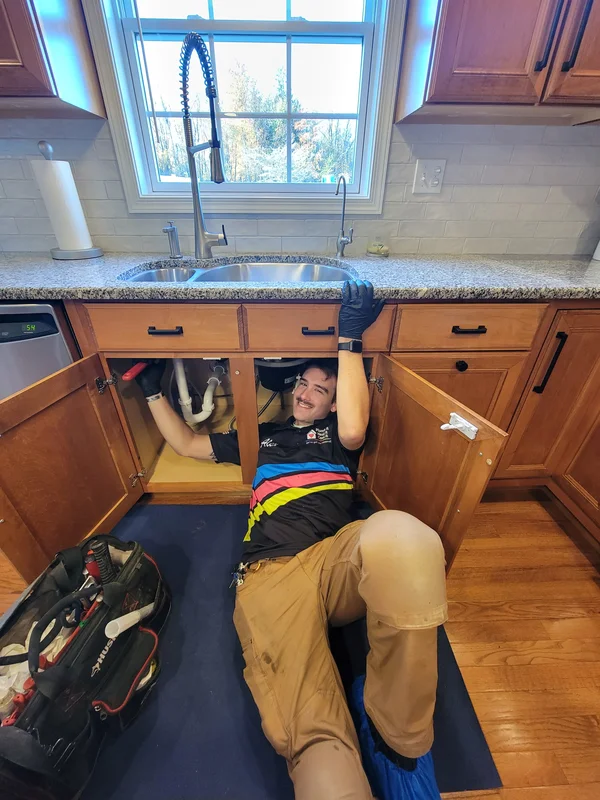 A repair technician lies on the floor under a kitchen sink, reaching inside the cabinet to work on plumbing, with tools nearby and sunlight coming through the window above.