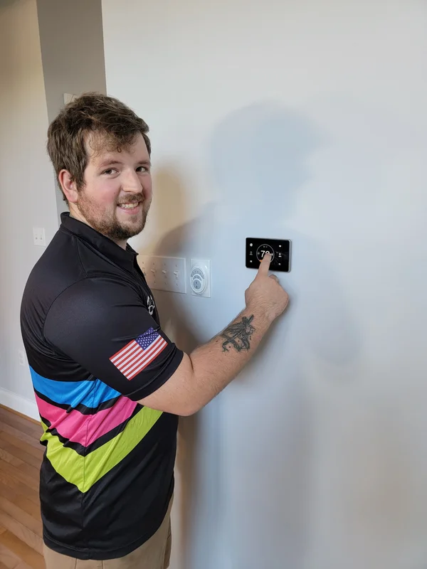 Man in a striped shirt is adjusting a thermostat on a wall, smiling at the camera.