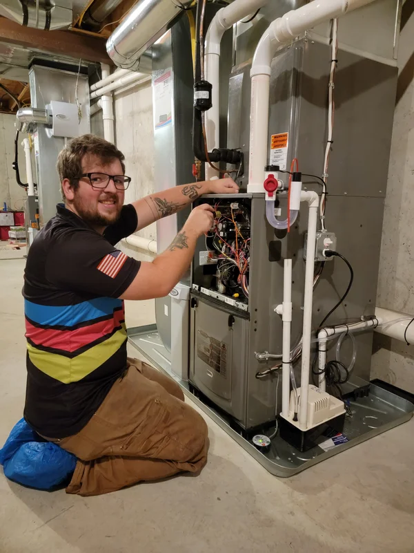 A man kneels on the floor and works on the wiring inside an open HVAC unit in a basement, using a screwdriver.