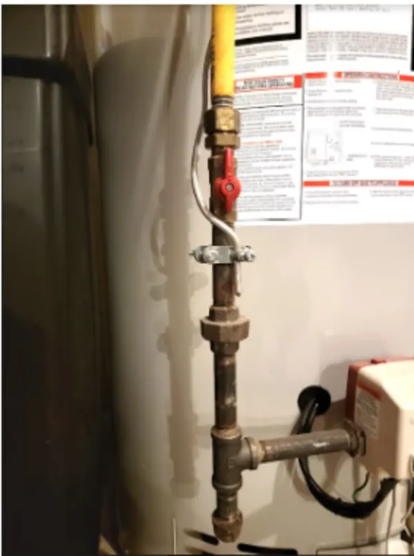 A close-up view of metal pipes and a yellow flexible gas line connected to a water heater, with instructions posted on the wall behind.