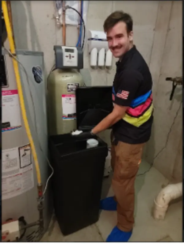 Person standing in a utility room, adding salt to a water softener unit while wearing brown pants, a striped shirt, and blue shoe covers.