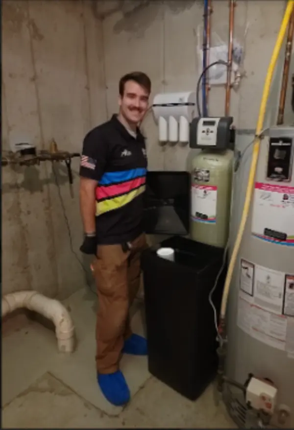 A person wearing gloves and blue shoe covers stands next to a water softener system in a basement with exposed pipes and concrete walls.
