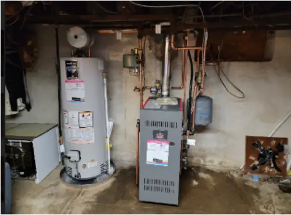 A residential basement with a water heater on the left and a gas furnace on the right, both installed against a concrete wall with exposed pipes and wiring.