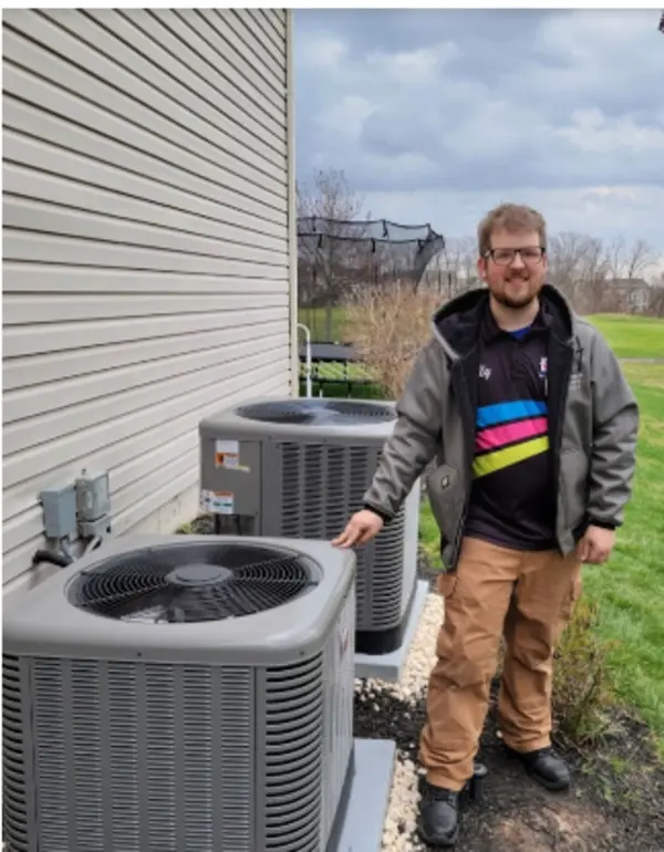 A man stands outside next to two large air conditioning units by the side of a house on a cloudy day.