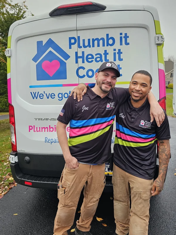 Two plumbers in matching uniforms stand in front of a company van with "Plumb it Heat it Cool it" and a house logo printed on the back.
