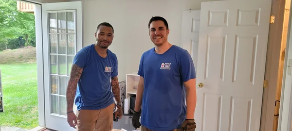 Two men wearing blue shirts stand inside a room under renovation, with an open door to the outside and tools visible nearby.